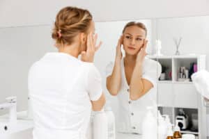 A young beautiful woman looking in the mirror and pulling the skin on her temples in relation to facelift surgery.