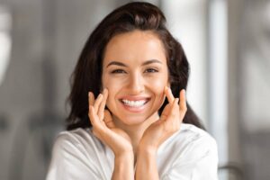 A woman with a cheerful smile and radiant skin gently touches her plump, youthful cheeks against a softly blurred, neutral background.