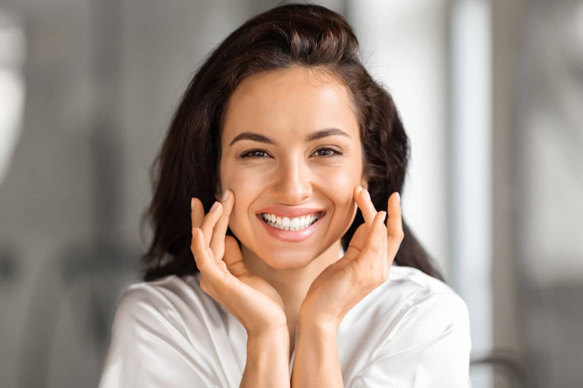 A woman with a cheerful smile and radiant skin gently touches her plump, youthful cheeks against a softly blurred, neutral background.