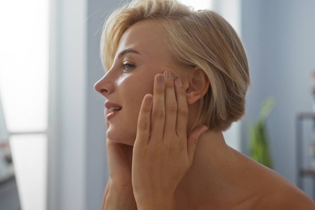 A blonde woman is shown in profile, delicately holding the base of her sharply defined jawline, emphasizing sculpted and volumetric facial contours.