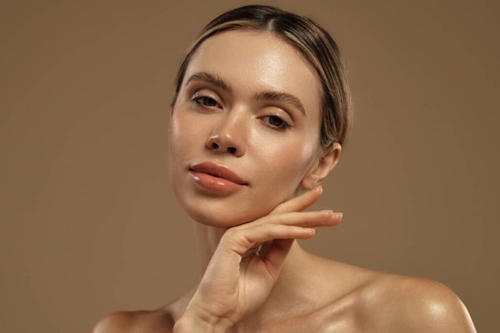 A woman with her hair pulled into a low bun poses with her hand under her chin, showcasing a smooth and balanced overall facial harmony.