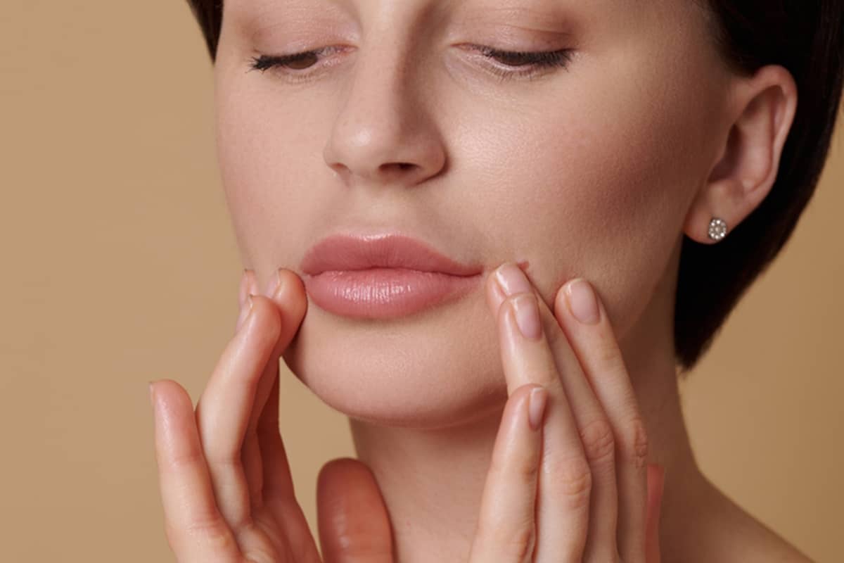 A close-up view of a woman’s lips as she uses her fingertips to carefully touch her softly enhanced, natural-looking pucker.