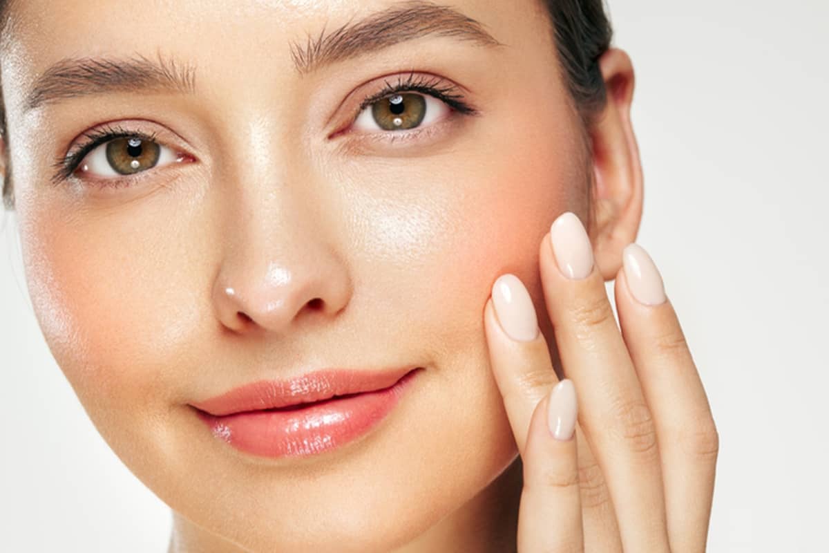 A woman with light-colored, neatly manicured oval nails touches her glowing cheek, displaying very smooth, even skin texture and brightness.