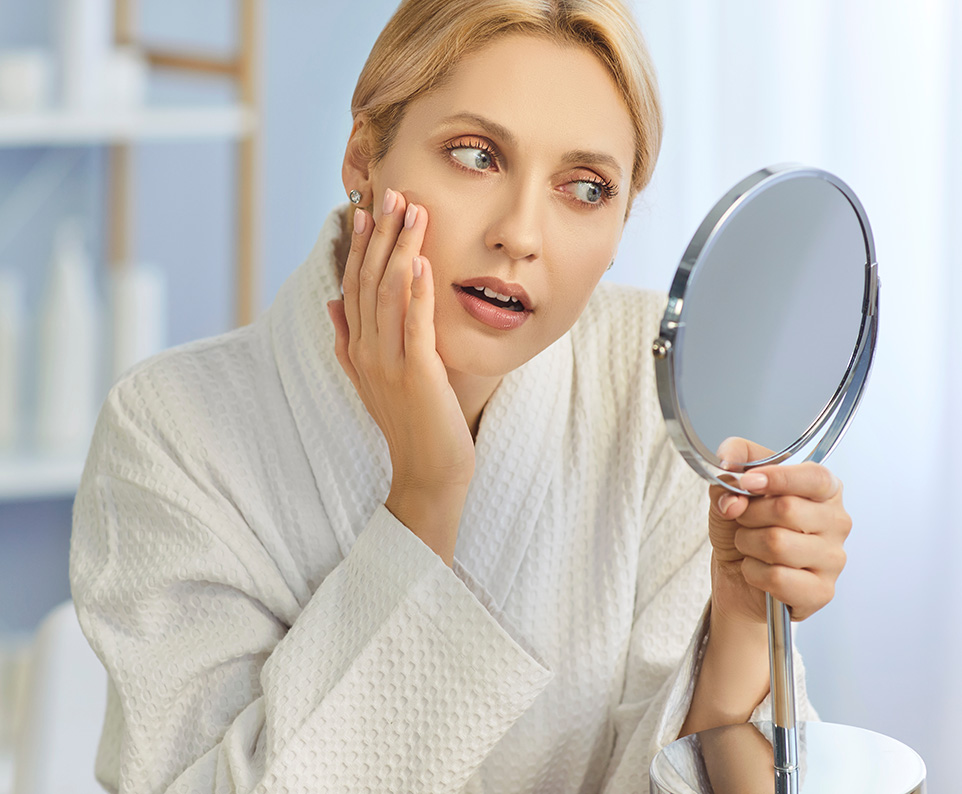 Woman examining cheeks in mirror illustrating candidacy for buccal fat removal and facial slimming.