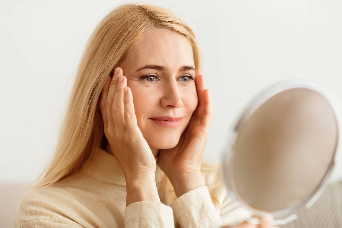 A blonde woman looking into a handheld mirror and using both hands to gently lift the skin on her cheeks and temples.