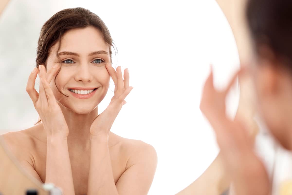 A smiling woman looking at her reflection in a circular mirror while gently touching the skin at the outer corners of her eyes.