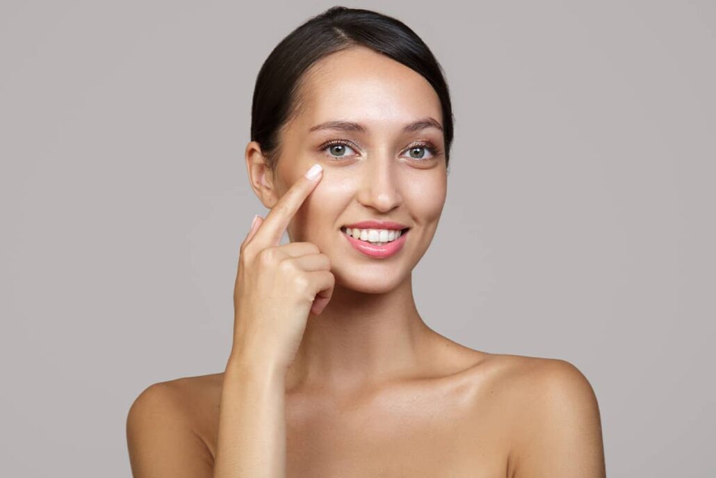 A smiling woman pointing a finger directly toward her lower eyelid to highlight the area where under-eye bags or fat deposits typically form.